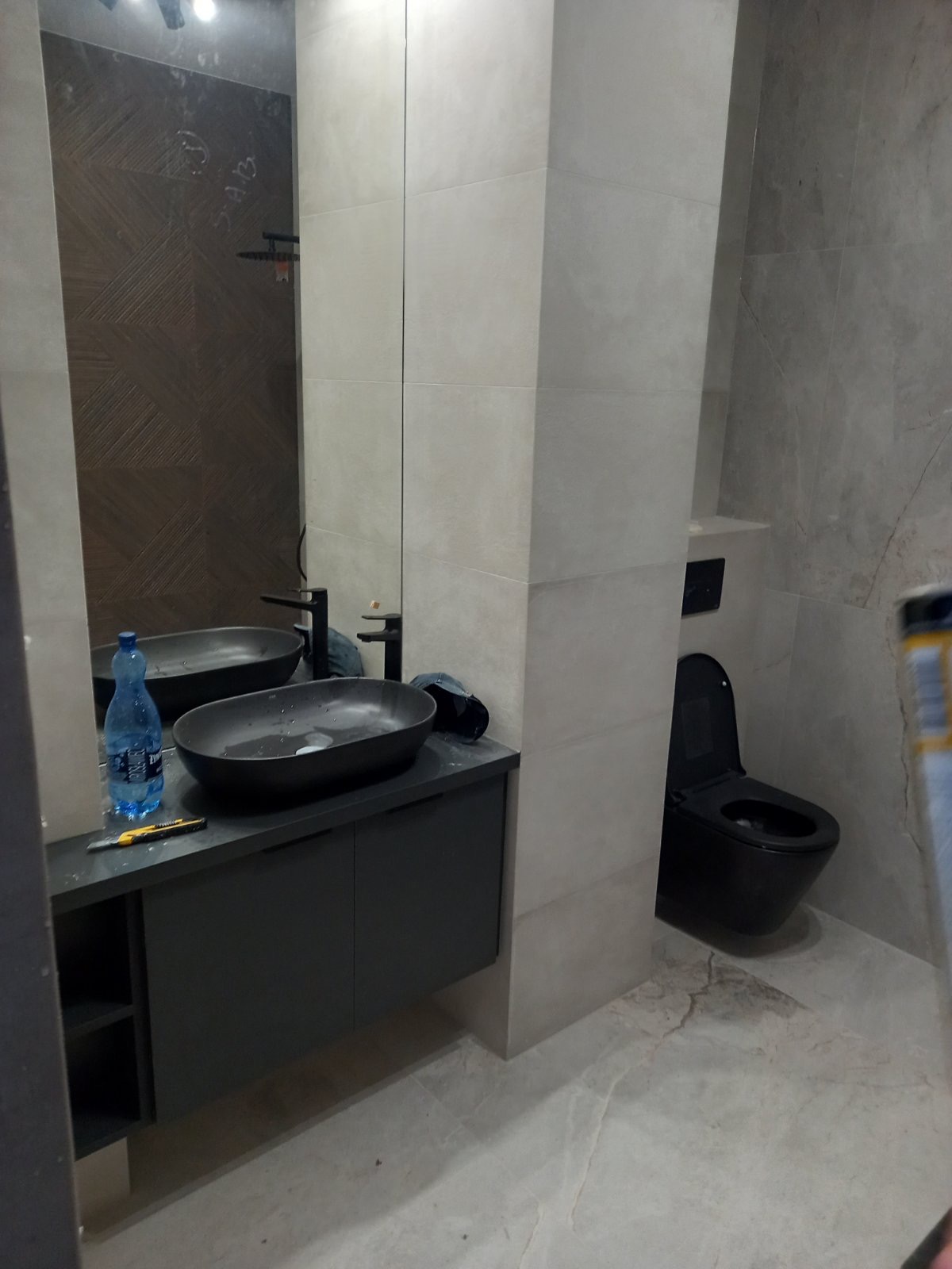 Nowoczesna łazienka z umywalką nablatową w kolorze czarnym, minimalistyczną armaturą i płytkami imitującymi marmur na ścianach oraz podłodze. Na blacie widoczny nóż do tapet oraz butelka wody.