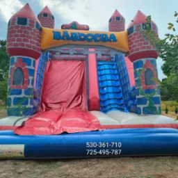 Kolorowy, dmuchany zamek z motywem średniowiecznym, zjeżdżalnią i schodami linowymi, ustawiony na trawniku, z widocznym numerem telefonu na niebieskiej podstawie.