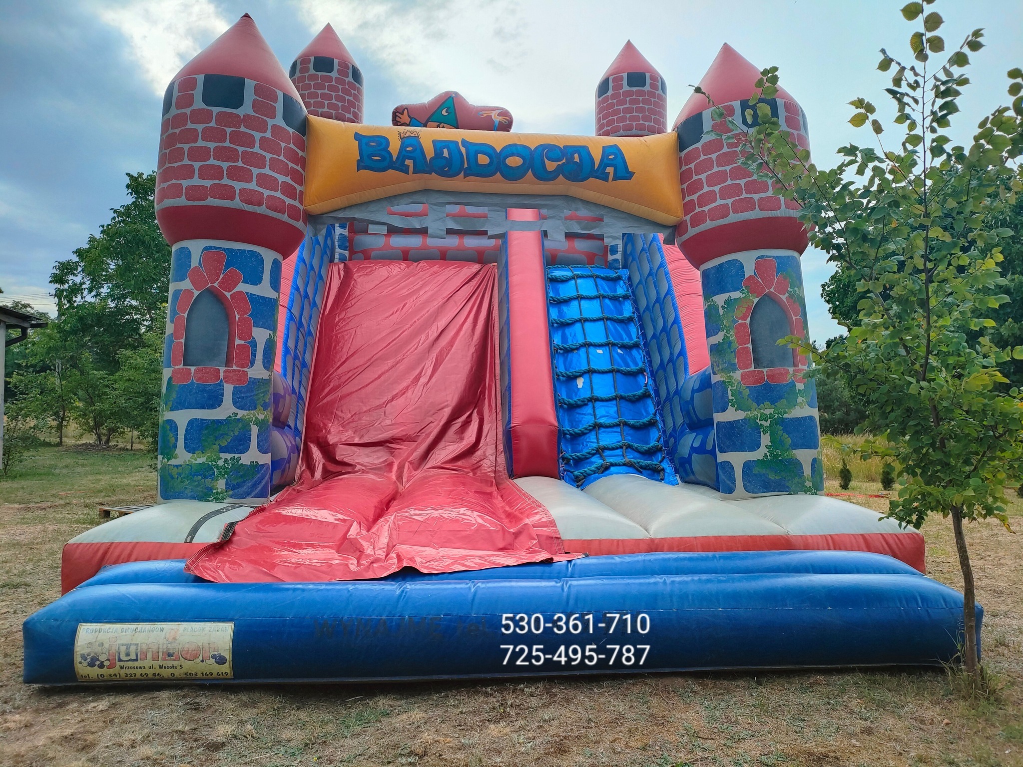 Kolorowy, dmuchany zamek z motywem średniowiecznym, zjeżdżalnią i schodami linowymi, ustawiony na trawniku, z widocznym numerem telefonu na niebieskiej podstawie.