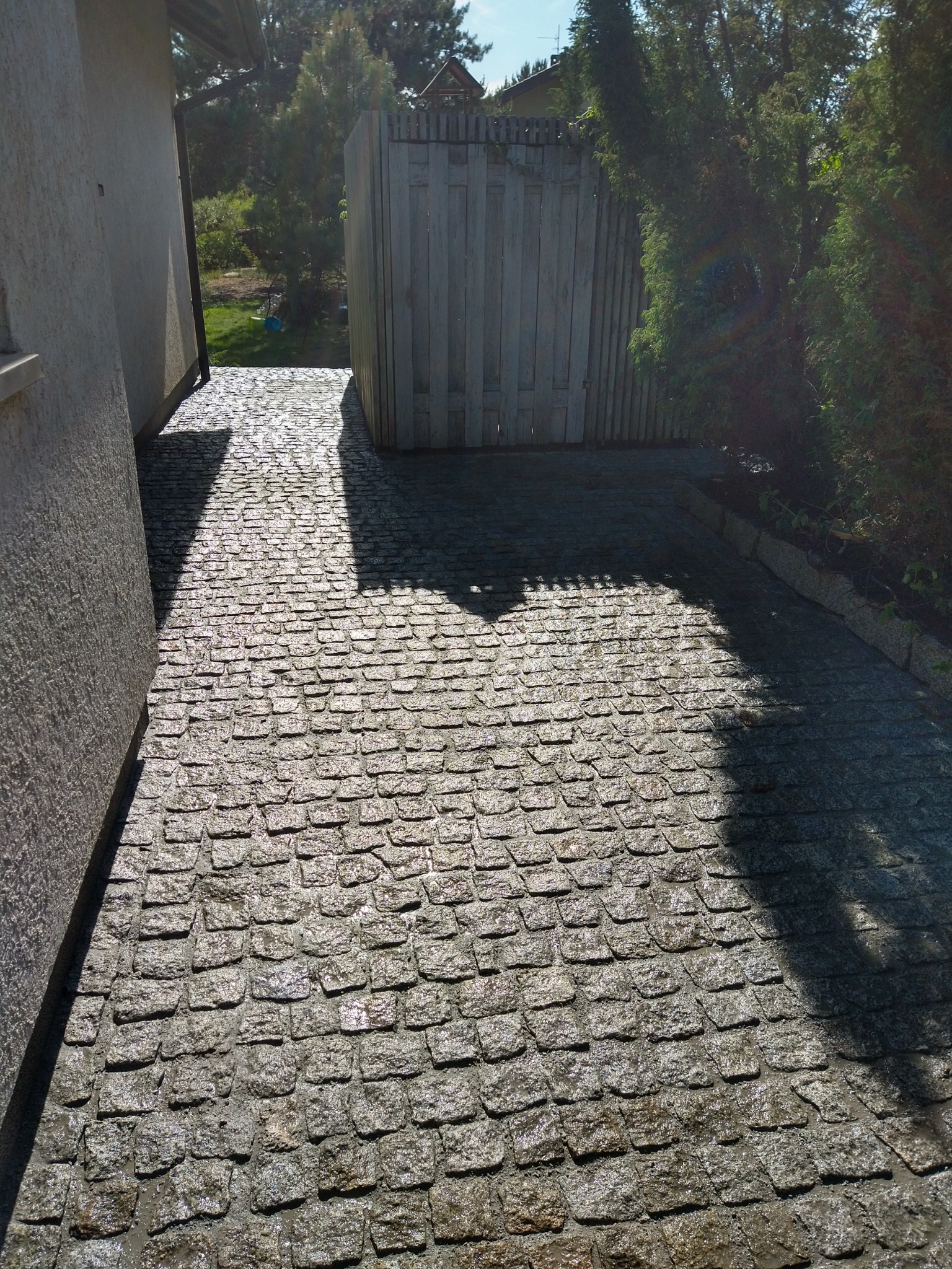 Świeżo ułożona, wilgotna nawierzchnia z kostki granitowej w odcieniach szarości, prowadząca wzdłuż ściany budynku i drewnianego ogrodzenia, z fragmentami cienia drzew.