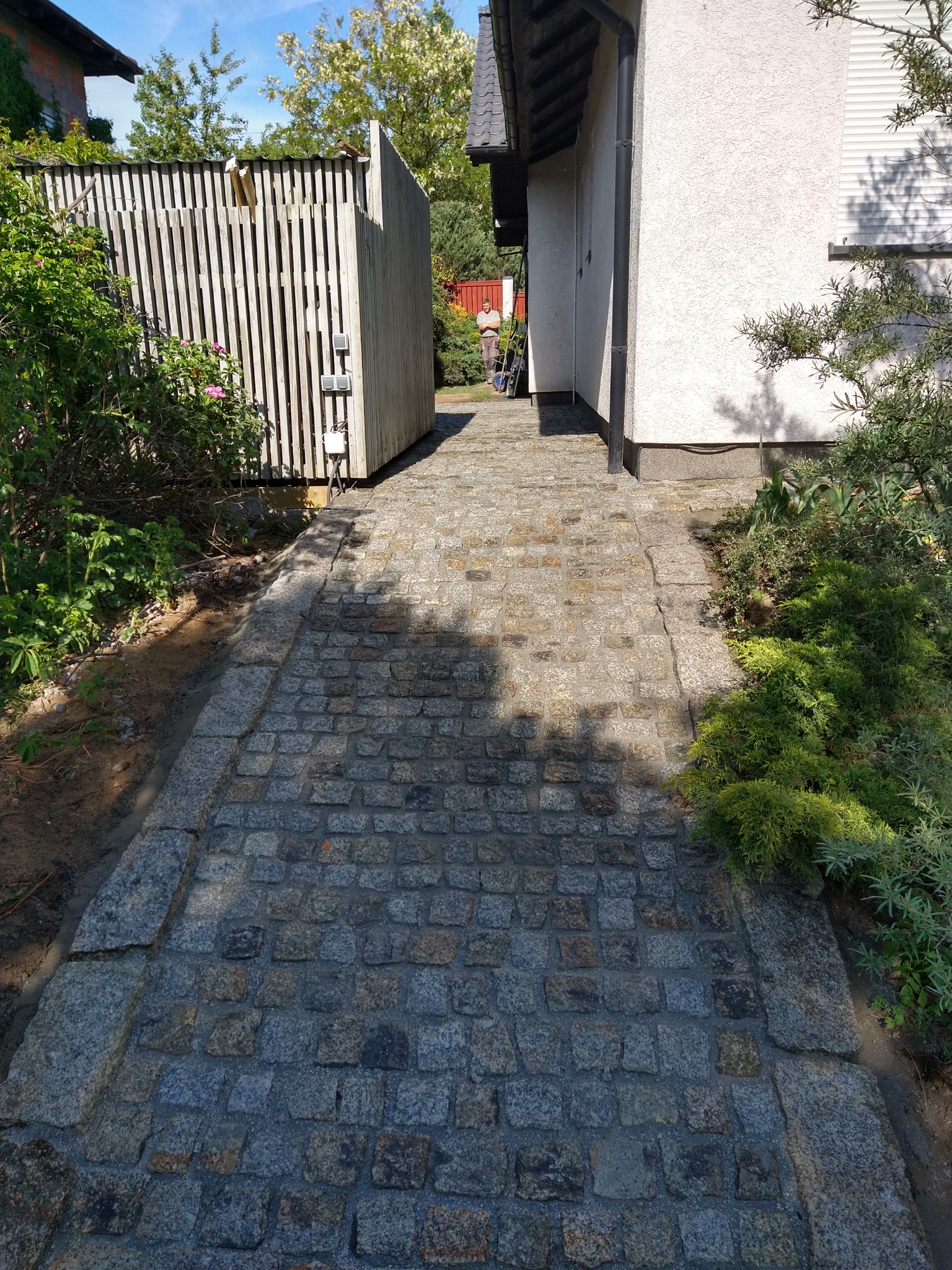 Granitowa ścieżka prowadząca wzdłuż budynku, z widocznym fragmentem drewnianego ogrodzenia i postacią w tle, otoczona zielenią.