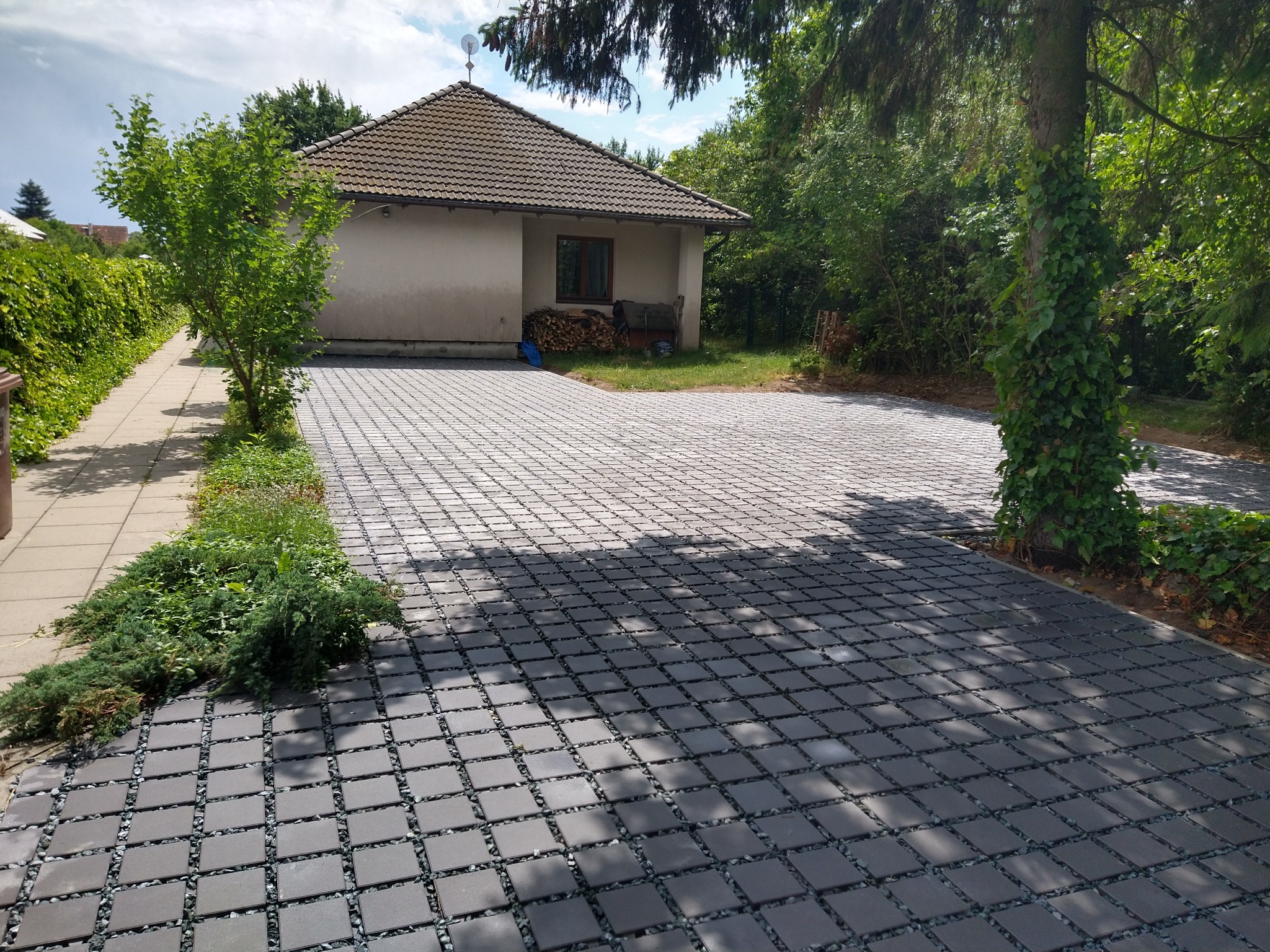 Szeroki podjazd wyłożony szarą kostką ażurową z wypełnieniem żwirowym, prowadzący do parterowego domu z jasną elewacją i stertą drewna przy ścianie. Fragment drzewa oplecionego bluszczem po prawej...