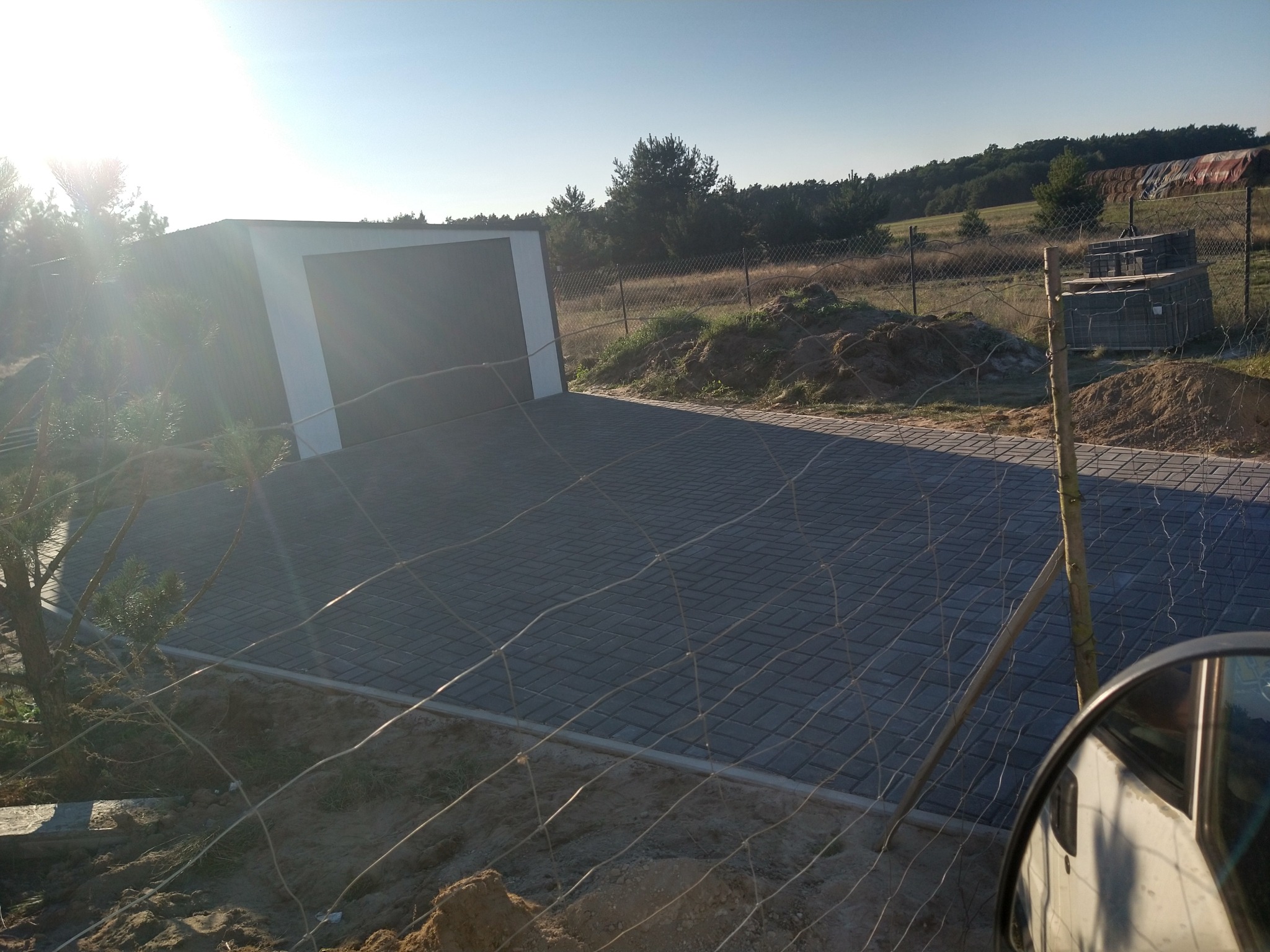 Ułożona, szara kostka brukowa przed garażem, widoczna przez siatkę ogrodzeniową. W tle drzewa i niebo.