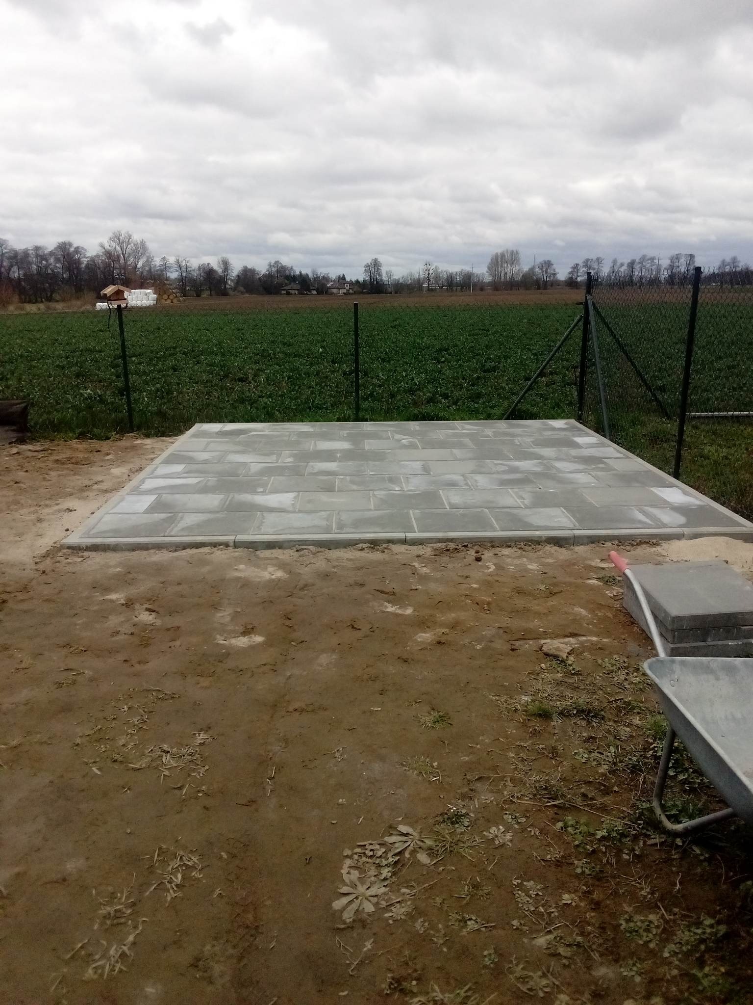 Ułożona prostokątna nawierzchnia z szarej kostki brukowej z obrzeżami na ziemi, widoczne taczki i pole uprawne w tle.