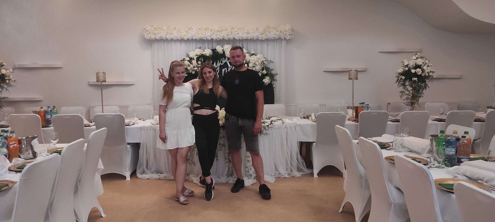 Trzy osoby pozują na tle udekorowanej sali weselnej z białymi kwiatami i stołami z białymi obrusami.
