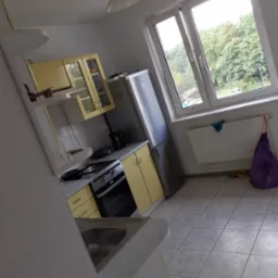 Jasna kuchnia z żółtymi szafkami, widok na okno i grzejnik z torbą obok, perspektywa z boku.