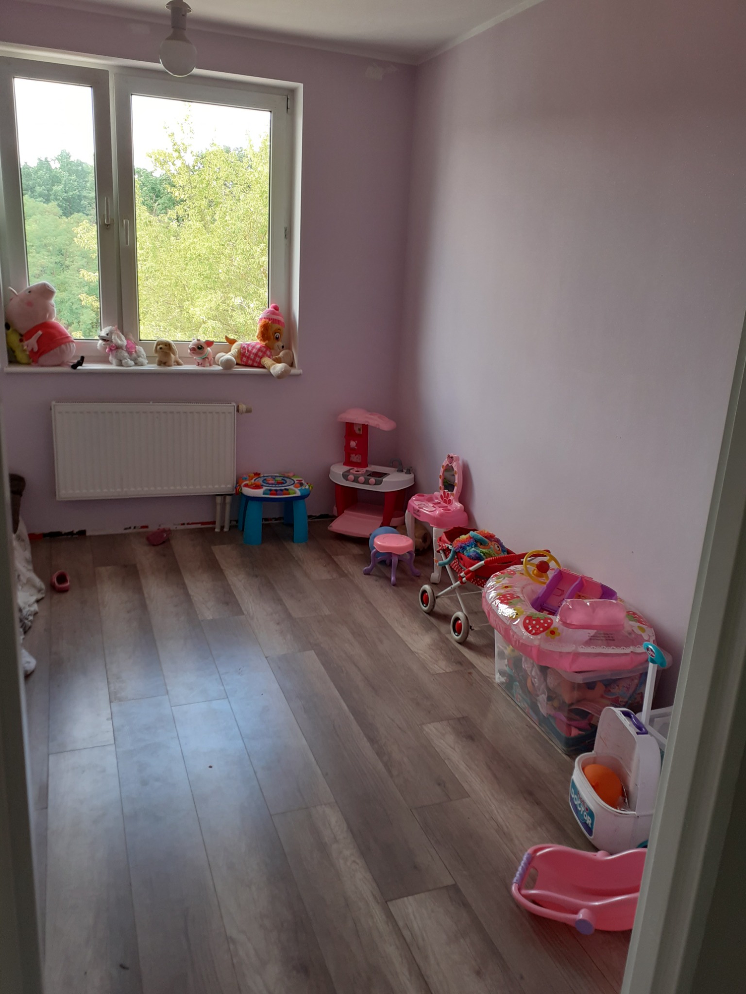 Pomalowany na liliowo pokój dziecięcy z zabawkami, widoczne ślady po taśmie malarskiej przy podłodze, okno z pluszakami na parapecie.