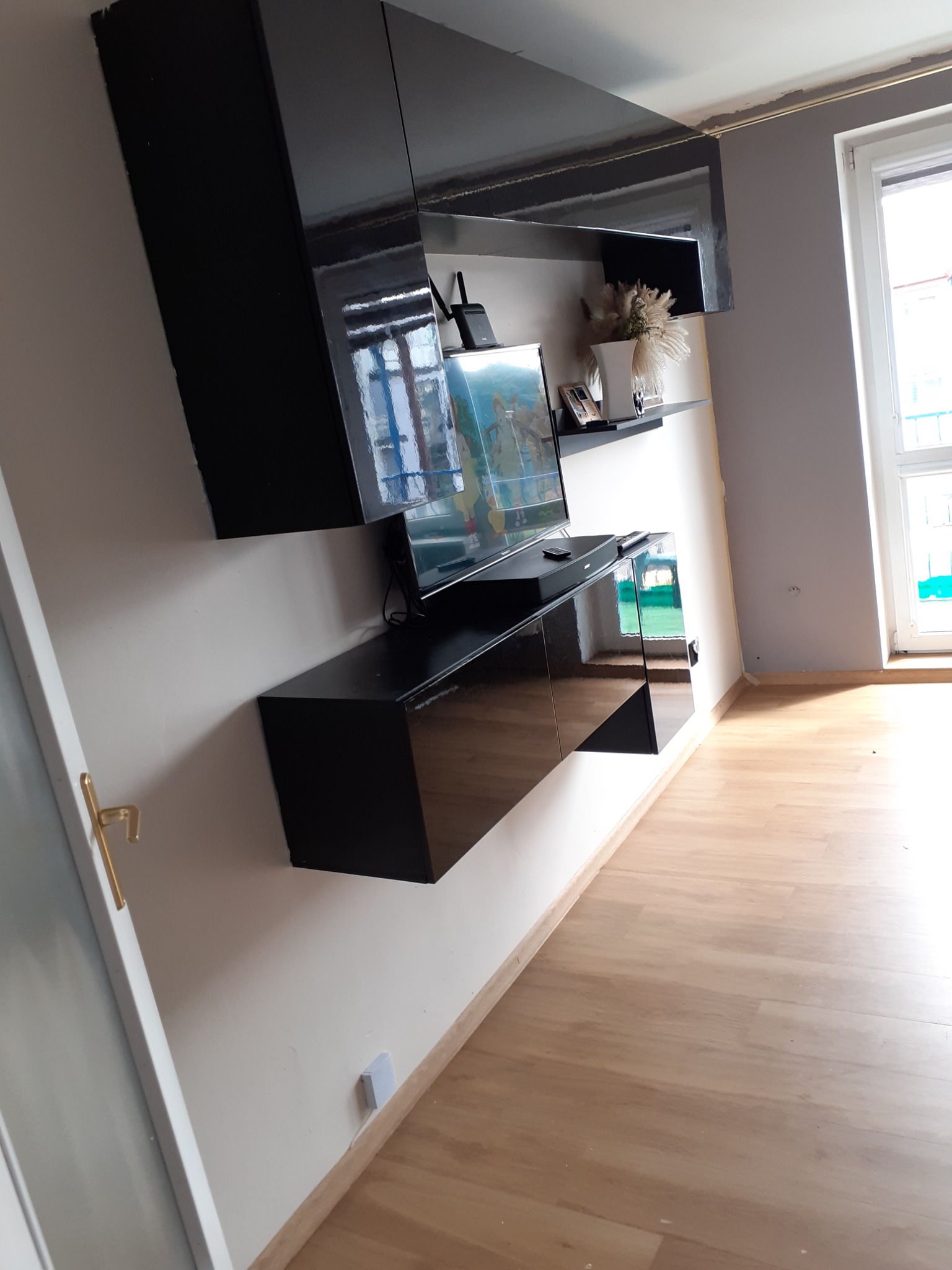 Czarne, lakierowane meble wiszące w salonie z telewizorem i dekoracjami, jasna podłoga z paneli, ściany w odcieniach bieli i szarości, widoczne ślady po naprawach na suficie.