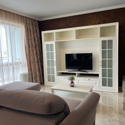 Revelo Wioletta Pietras - Jasne wnętrze salonu z beżową sofą, białą zabudową meblową z telewizorem i marmurową podłogą. Okno z dekoracyjnymi zasłonami w tle. Eleganckie i przestronne.