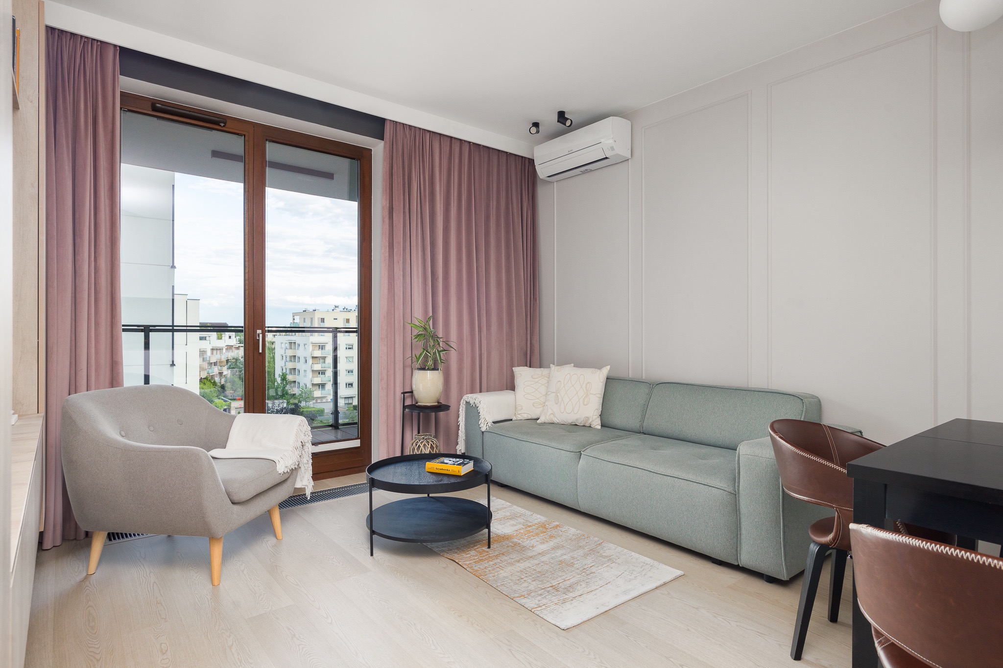 Jasne, nowocześnie urządzone wnętrze mieszkania z szarozieloną sofą, szarym fotelem z narzutą, okrągłym stolikiem kawowym i widokiem na miasto z balkonu.