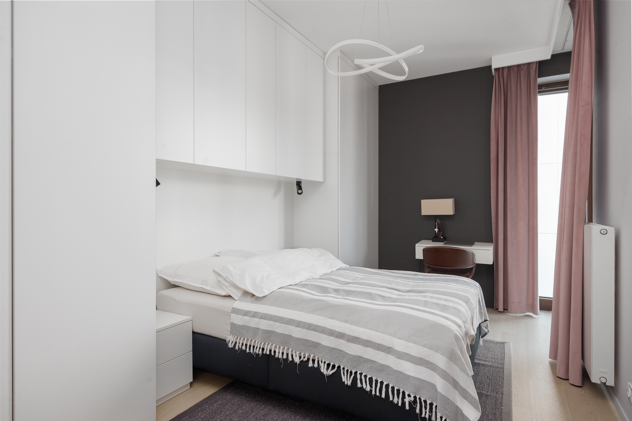 Nowoczesna sypialnia z łóżkiem, biurkiem i minimalistyczną lampą wiszącą, utrzymana w bieli i szarościach z akcentami brudnego różu na zasłonach.