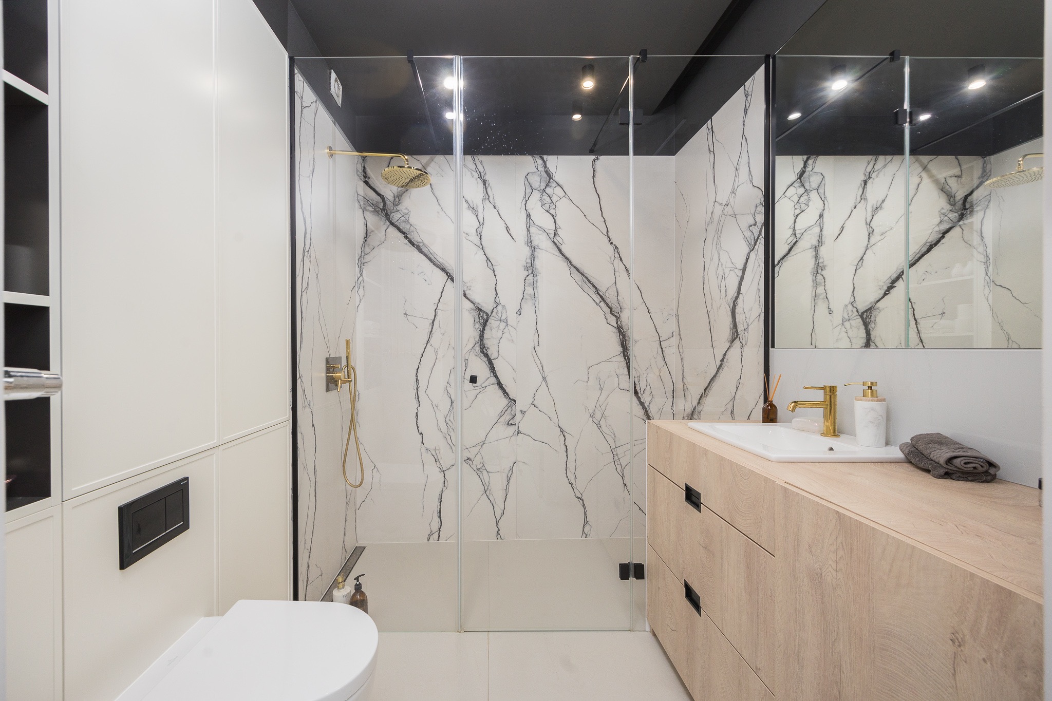 Nowoczesna łazienka z kabiną prysznicową wykończoną płytkami imitującymi marmur, złotą armaturą i drewnianą szafką pod umywalką.