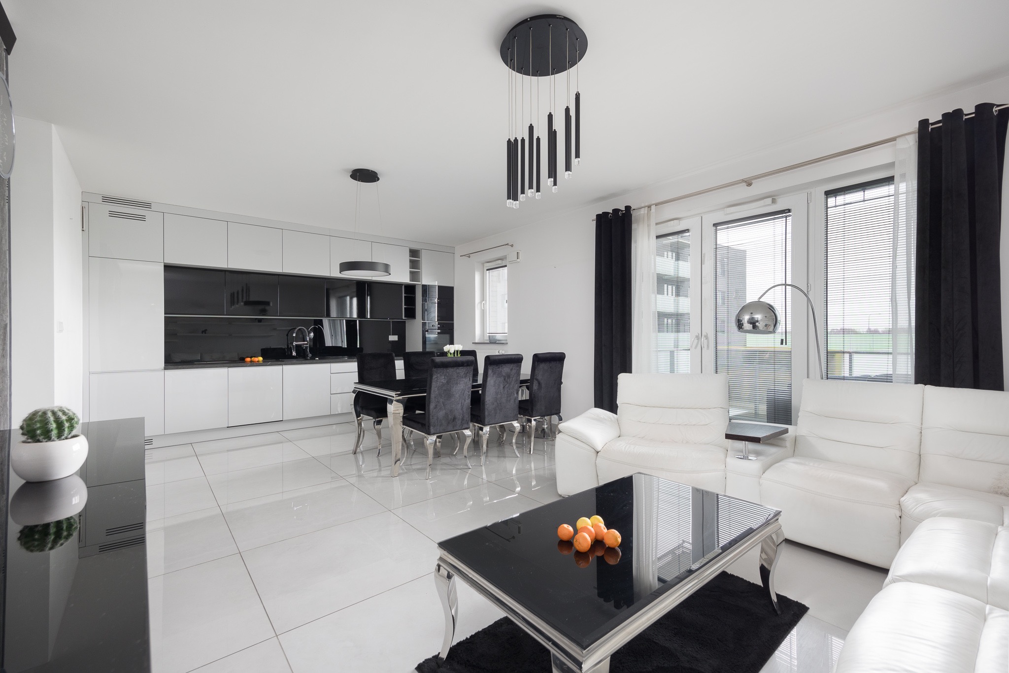 Nowoczesne wnętrze: biała skórzana sofa, czarny stolik kawowy z owocami, stół z krzesłami, kuchnia z czarno-białymi szafkami, białe płytki podłogowe z połyskiem, czarne zasłony i lampa podłogowa.