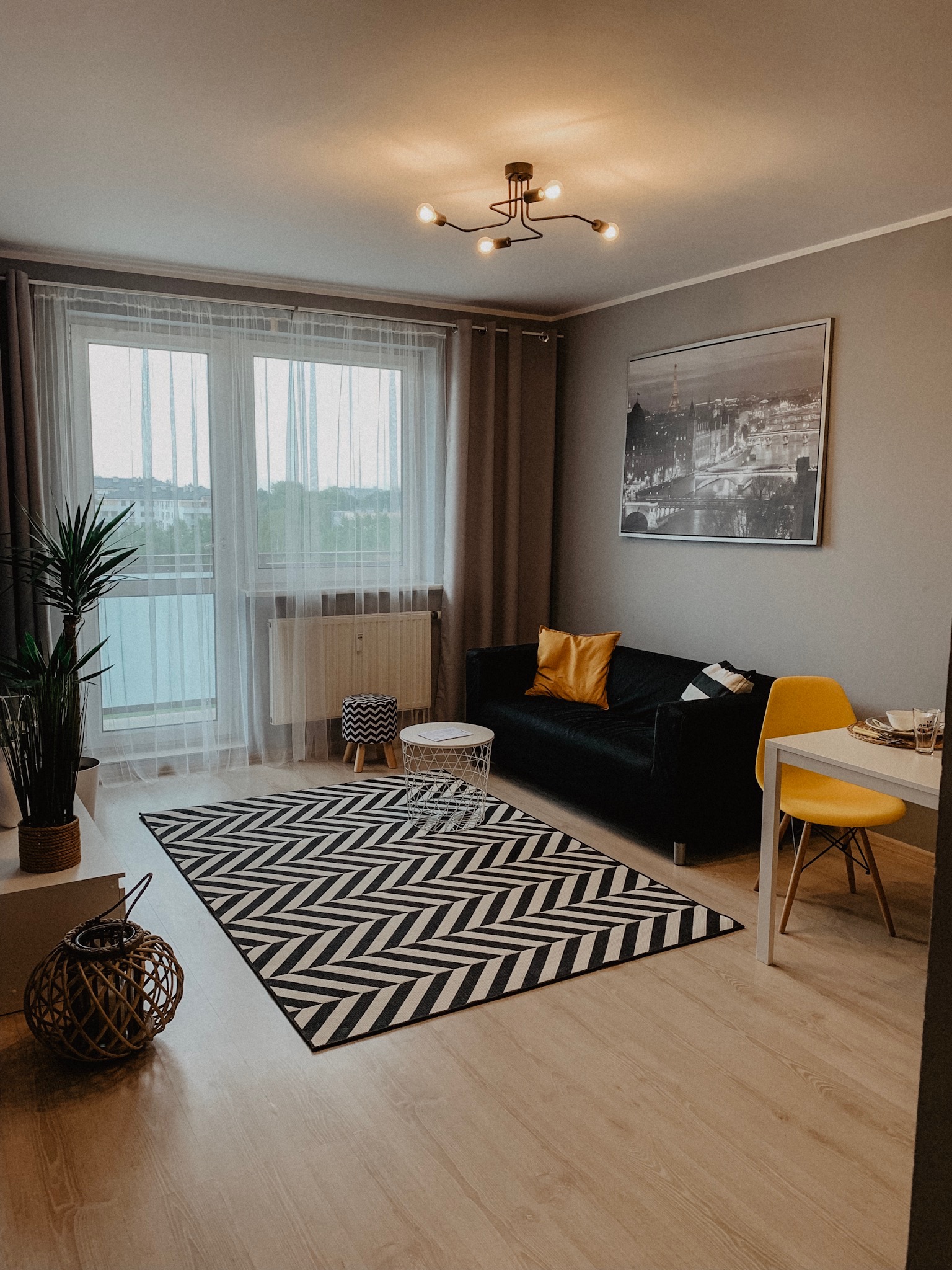 Jasne wnętrze mieszkania z czarną sofą, żółtym krzesłem i wzorzystym dywanem. Na ścianie grafika z motywem miejskim. Widok z okna na zieleń.