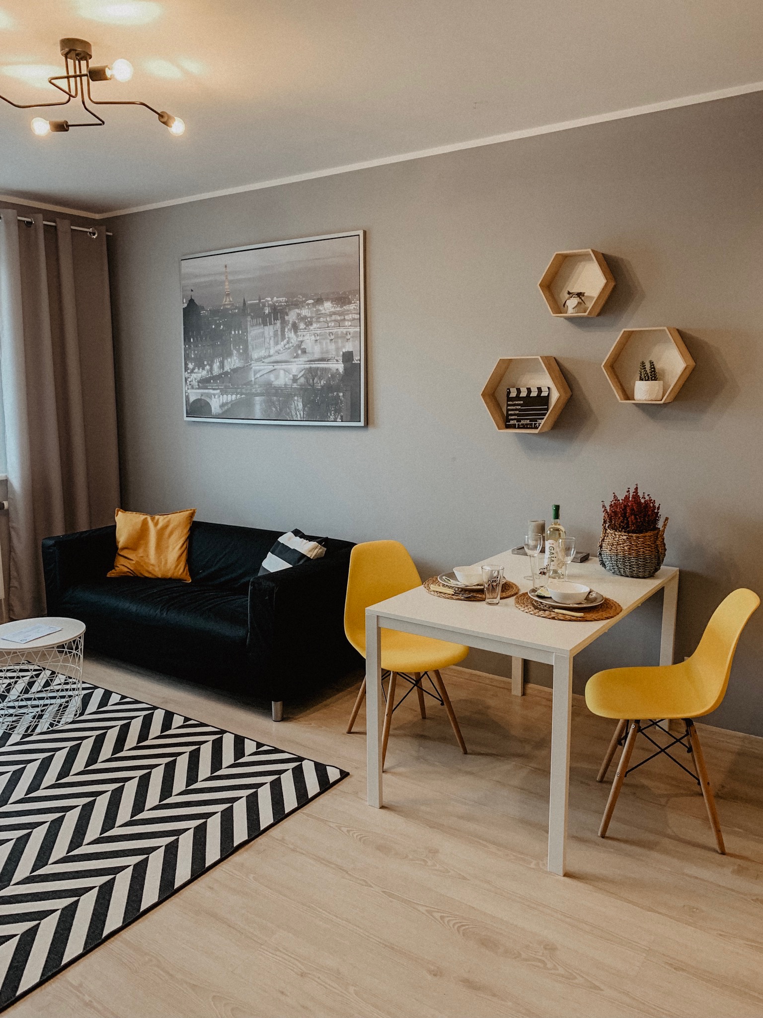 Jasne wnętrze mieszkania z czarną sofą, żółtymi krzesłami i stołem z zastawą. Na ścianie grafika z wieżą Eiffla i heksagonalne półki. Nowoczesny design.