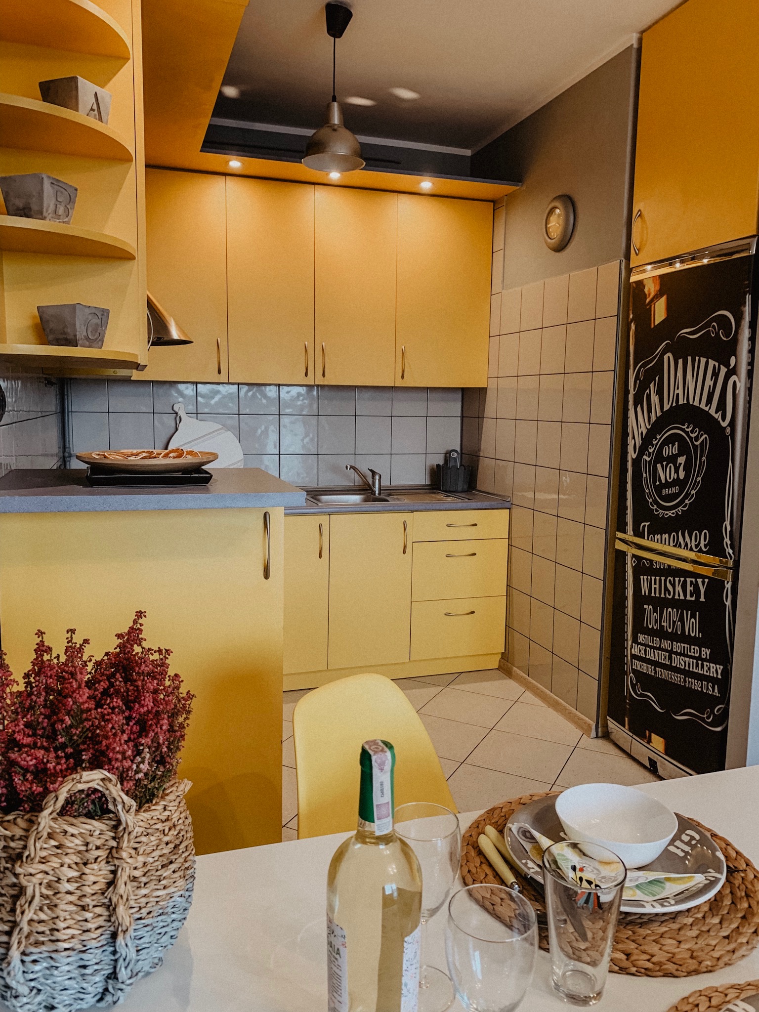 Żółta kuchnia z plakatem Jack Daniel's na lodówce, nakryty stół z kwiatami. Prezentacja wnętrza mieszkania na sprzedaż z akcentami dekoracyjnymi.