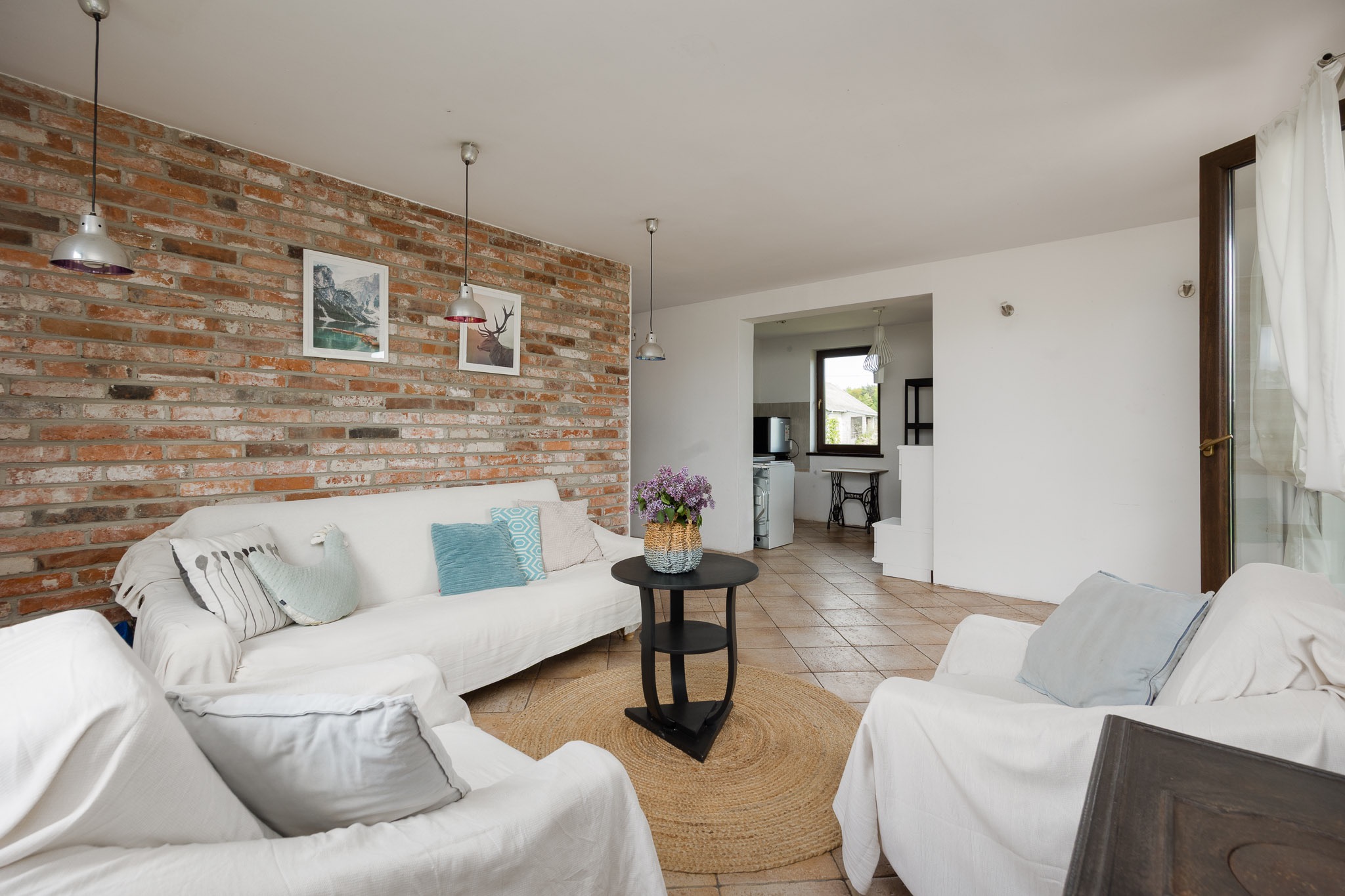 Jasne wnętrze: ceglana ściana z obrazami, białe meble, okrągły dywan i kwiaty w koszyku. Widok na kuchnię z oknem i maszyną do szycia. Sprzedaż mieszkania.