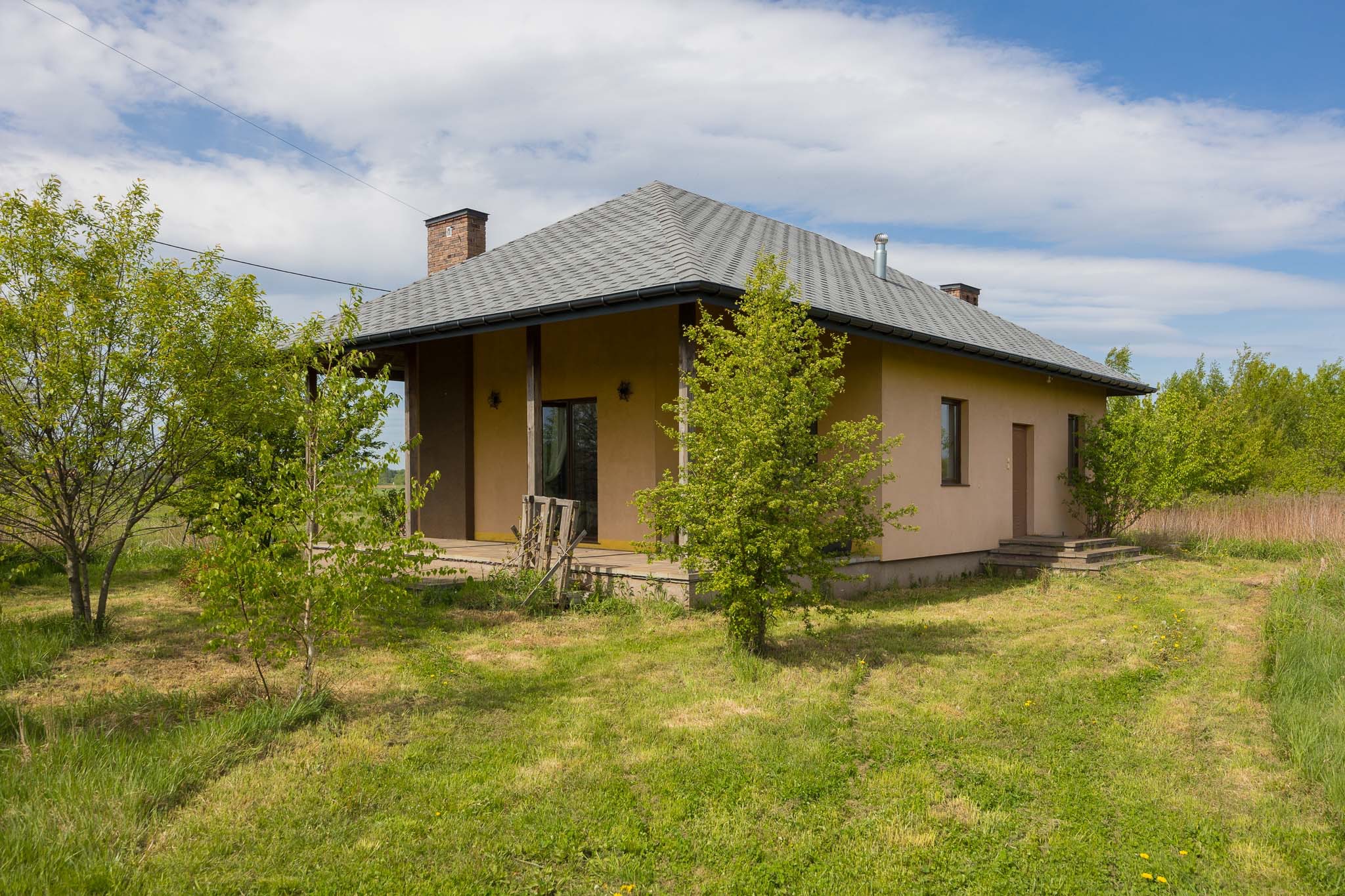 Parterowy dom z werandą w otoczeniu zieleni, na tle błękitnego nieba. Dach pokryty szarą dachówką, elewacja w odcieniach beżu. Widoczne drzewa i trawnik.