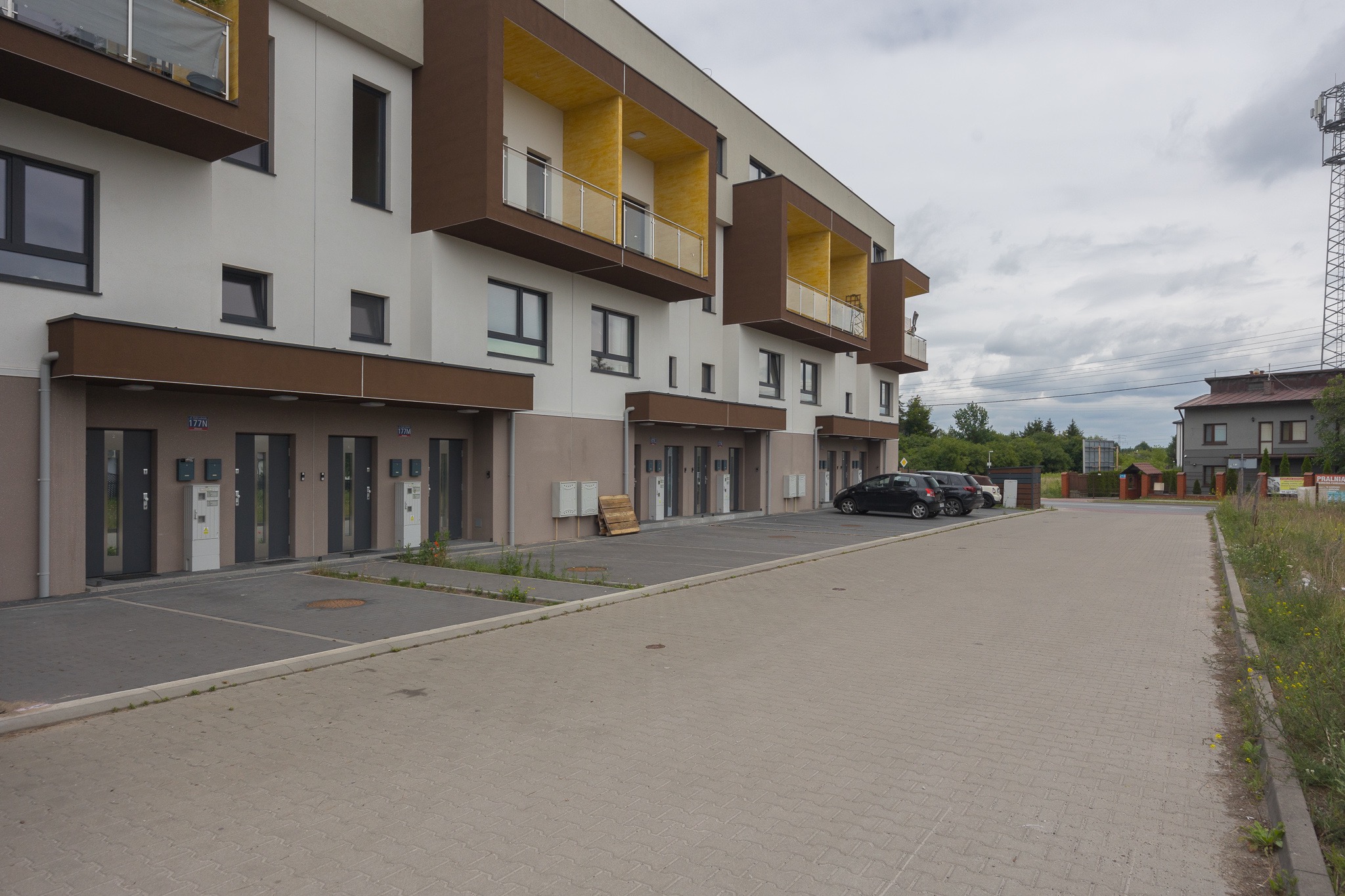 Nowoczesny budynek apartamentowy z balkonami w odcieniach brązu i beżu, z miejscami parkingowymi i chodnikiem z kostki brukowej. Widok z zewnątrz, pochmurne niebo.