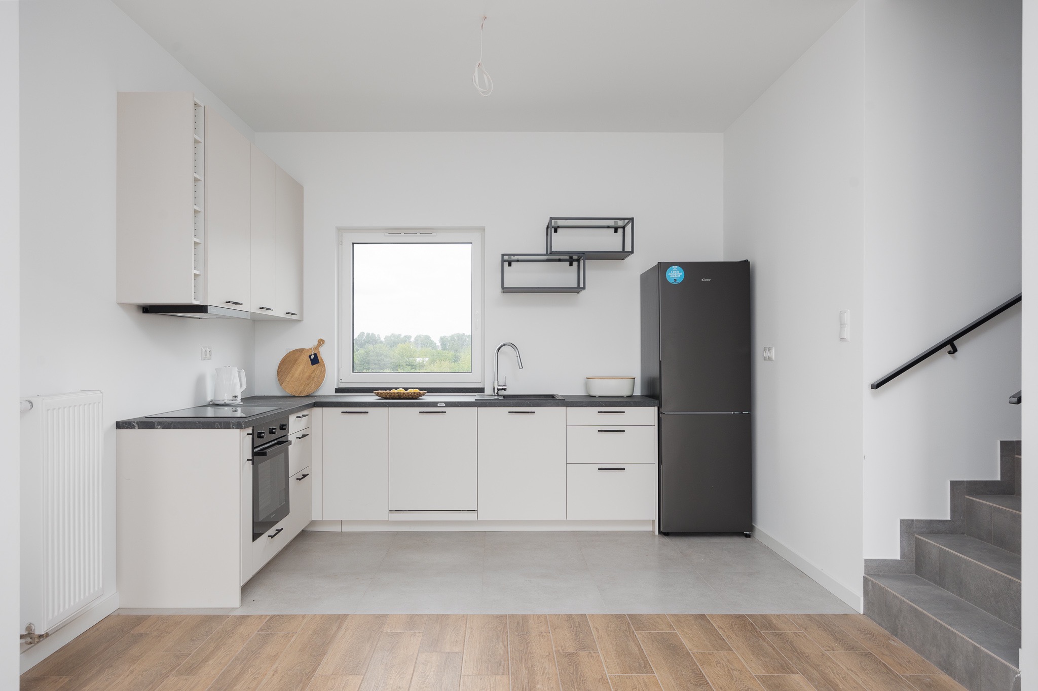 Jasna, minimalistyczna kuchnia z czarnymi akcentami: czarny blat, lodówka i półki. Drewniana podłoga przechodzi w szare płytki. Schody prowadzą na górę.
