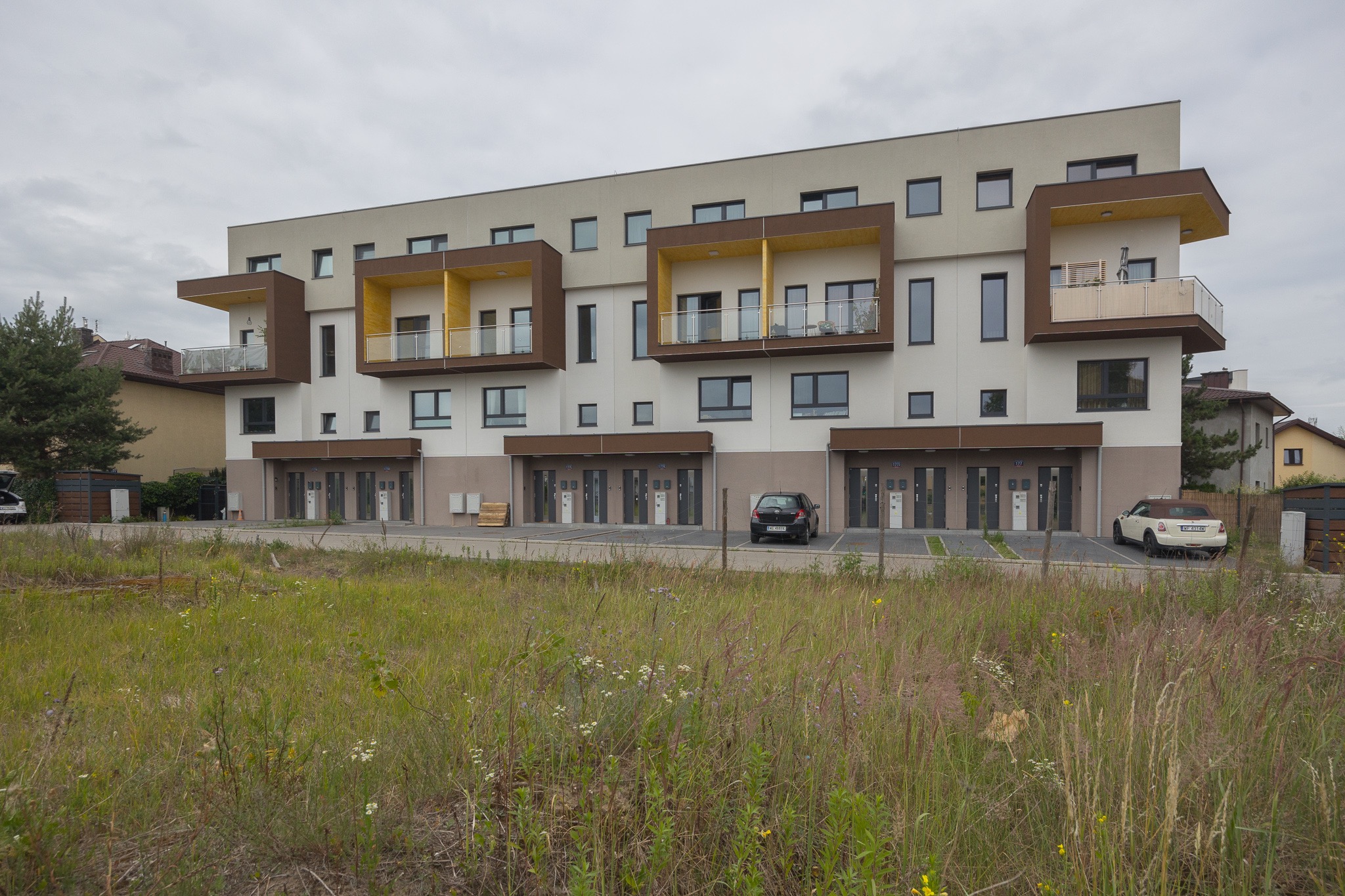 Nowoczesny budynek wielorodzinny z balkonami w odcieniach brązu i żółci, otoczony zielenią. Widoczne zaparkowane samochody i skrzynki pocztowe przy wejściach.