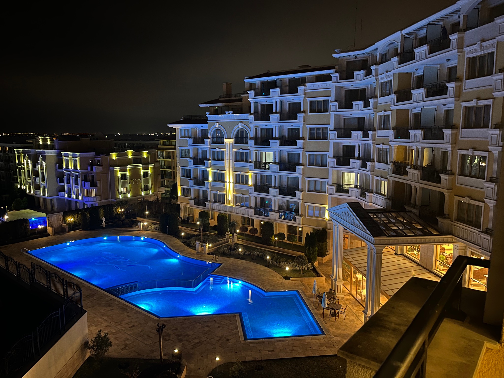 Nocny widok na luksusowy apartamentowiec z basenem oświetlonym na niebiesko, widok z balkonu. W tle widoczne światła miasta.