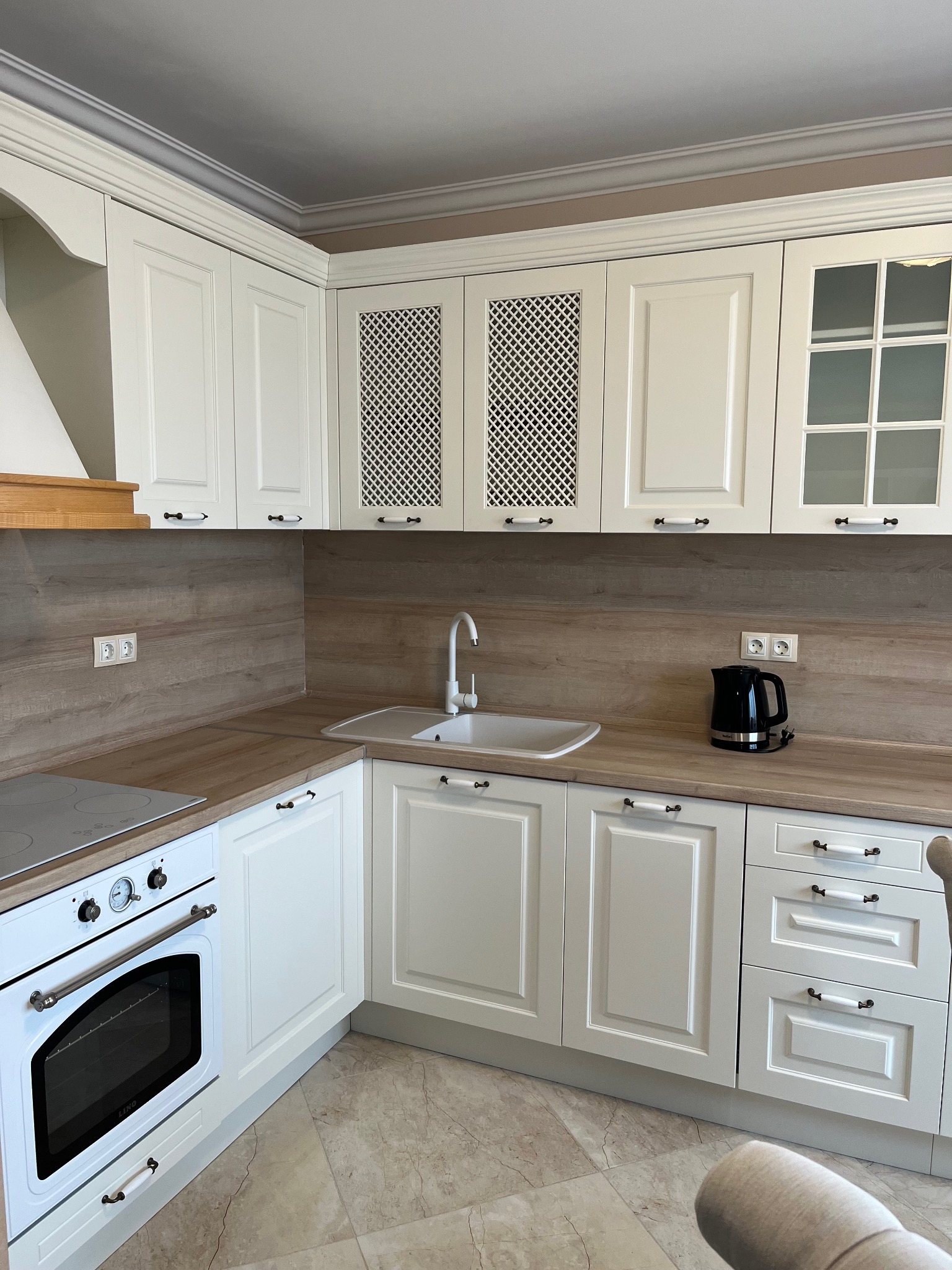 Jasna, klasyczna kuchnia z drewnianym blatem i białymi frontami szafek z ażurowymi wstawkami. Widoczny piekarnik, zlew i czajnik elektryczny.