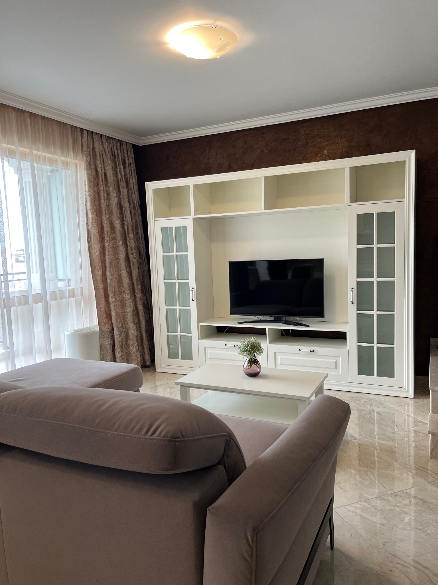 Jasne wnętrze salonu z beżową sofą, białą zabudową meblową z telewizorem i marmurową podłogą. Okno z dekoracyjnymi zasłonami w tle. Eleganckie i przestronne.