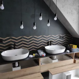 Dwie owalne umywalki nablatowe z czarną armaturą, zamontowane na szafce z drewnianym blatem w łazience na poddaszu; nad umywalkami ciemne płytki i dekoracyjny pas z wzorem w jodełkę, a nad nimi...