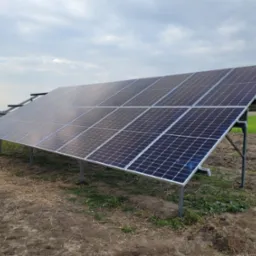 Instalacja fotowoltaiczna na polnej działce z widocznymi metalowymi wspornikami i panelami w ułożeniu rzędowym, tło: zielone pole i pochmurne niebo.