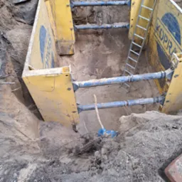 Wykonany wykop zabezpieczony żółtymi szalunkami stalowymi z drabinką, widoczne niebieskie rury wzmacniające konstrukcję, ziemia piaszczysta.