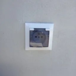 Gniazdko elektryczne zewnętrzne z klapką ochronną, zamontowane na szarej, teksturowanej ścianie. Klapka jest lekko uchylona, ukazując wnętrze gniazdka.