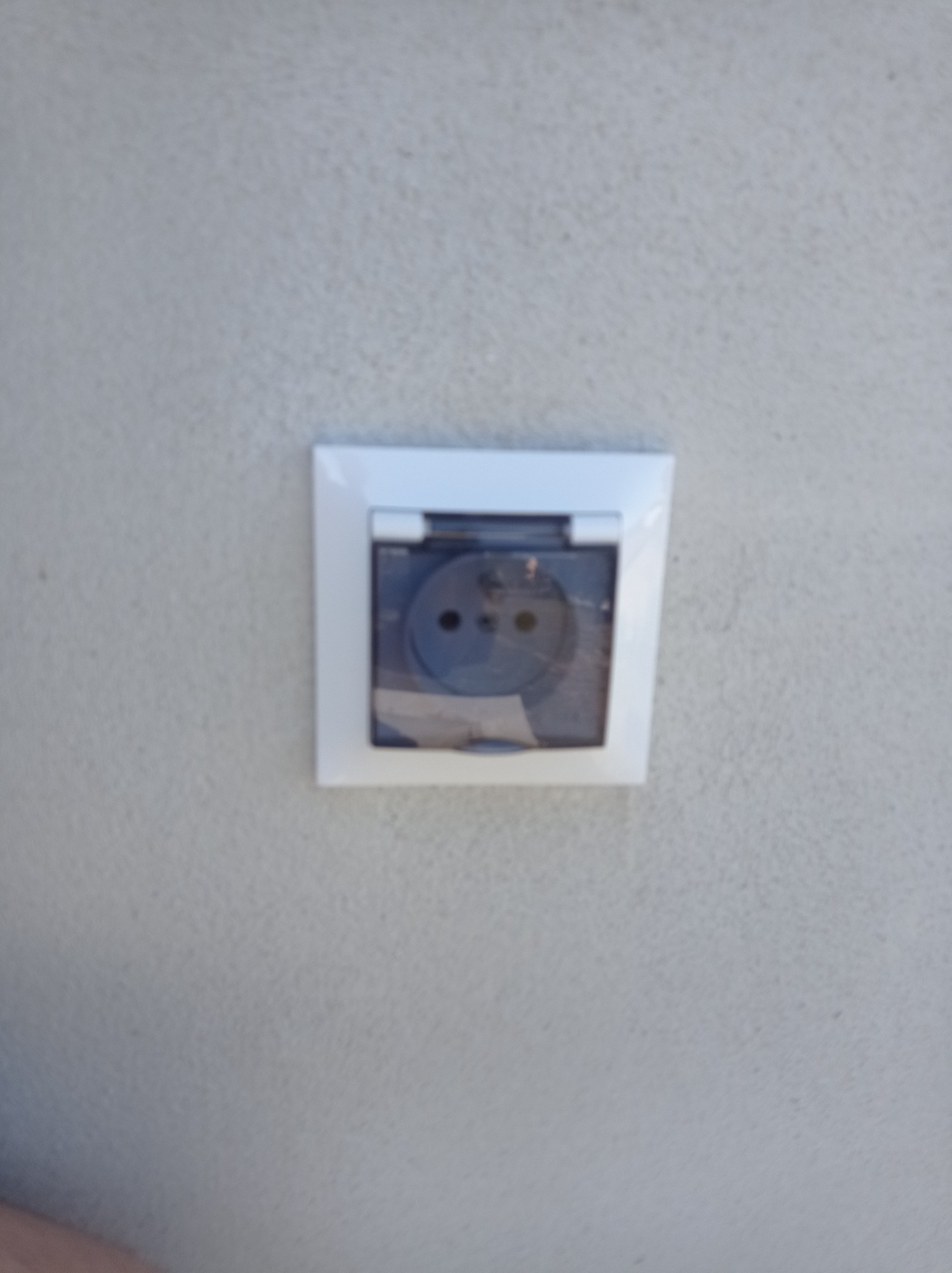 Gniazdko elektryczne zewnętrzne z klapką ochronną, zamontowane na szarej, teksturowanej ścianie. Klapka jest lekko uchylona, ukazując wnętrze gniazdka.