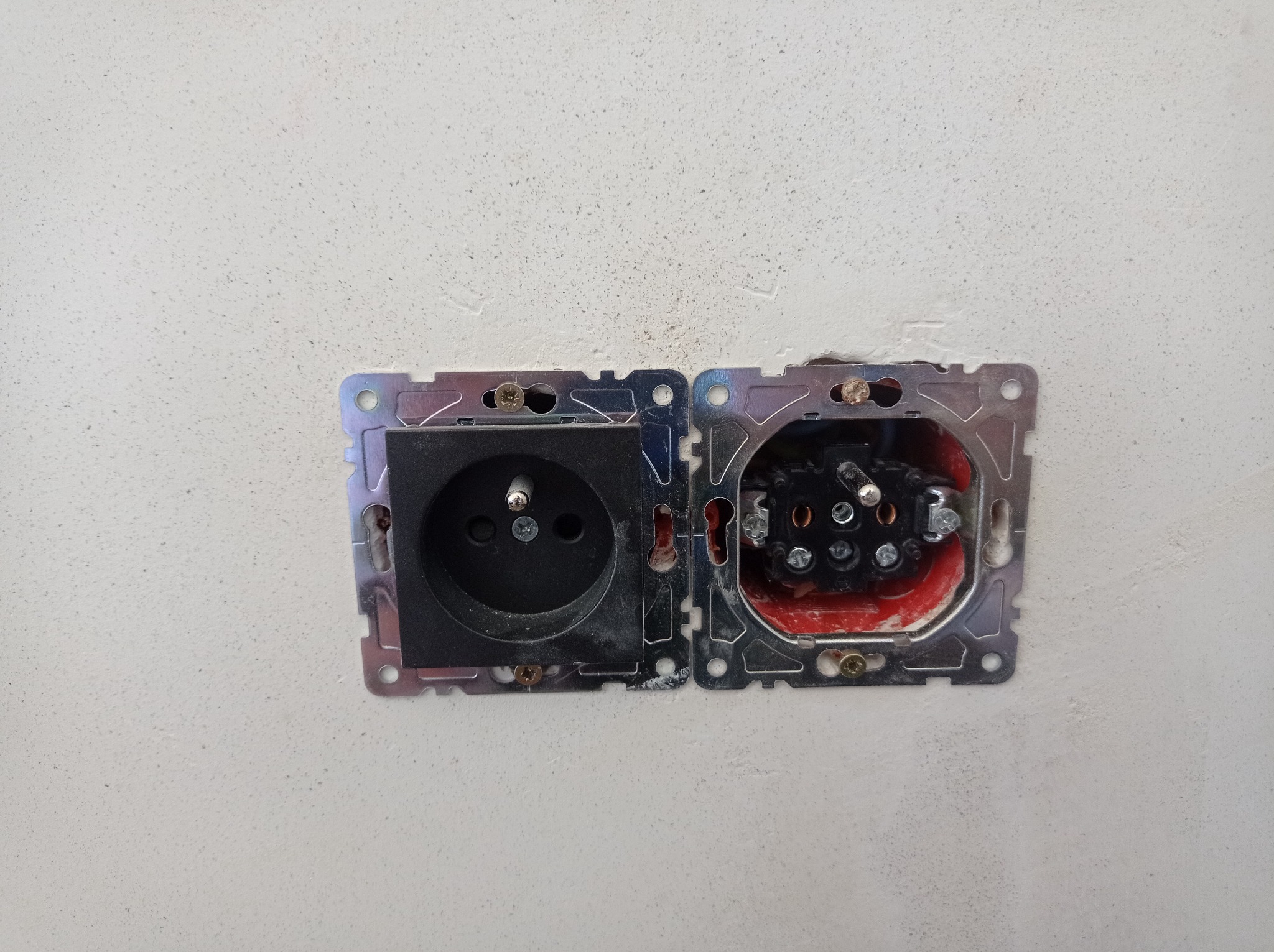 Podwójne gniazdko elektryczne w trakcie montażu w surowej, jasnej ścianie. Po lewej zamontowana czarna wtyczka, po prawej widoczne przewody w puszce, zabezpieczone czerwoną izolacją.