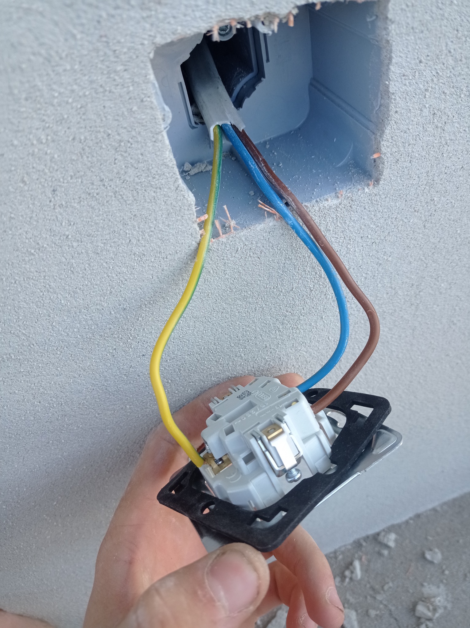 Podłączenie przewodów do gniazdka elektrycznego w surowej ścianie, widoczne przewody: żółto-zielony, niebieski i brązowy. Gniazdko trzymane w dłoni, gotowe do montażu w puszce.