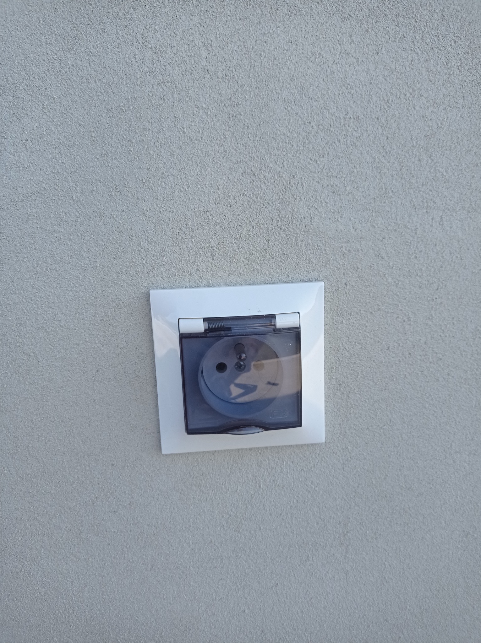 Zewnętrzne gniazdo elektryczne z białą ramką i przezroczystą klapką zabezpieczającą, zamontowane na szarej, chropowatej ścianie.