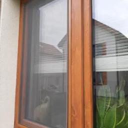 Brązowa rama okienna z zamontowaną siatką przeciw owadom; w tle widoczny kot siedzący na parapecie wewnętrznym.