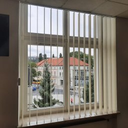 Problind Warszawa - Zamknięte, kremowe żaluzje pionowe w oknie biurowym z widokiem na plac miejski z budynkami i przechodniem.