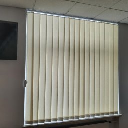 Problind Warszawa - Beżowe pionowe lamele zasłaniają okno w biurze, widoczny fragment ściany z telewizorem i tablicą suchościeralną.