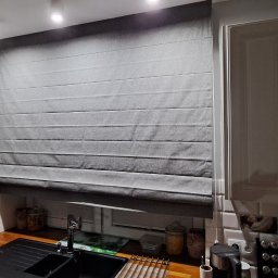 Problind Warszawa - Szara roleta rzymska zamontowana w kuchni nad zlewem, widoczne białe szafki kuchenne i dekoracje na blacie.