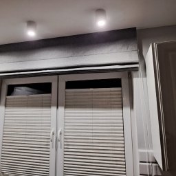 Problind Warszawa - Szara roleta rzymska zamontowana nad oknem z plisowanymi żaluzjami w kuchni z białymi szafkami.