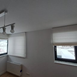 Problind Warszawa - Jasne, minimalistyczne wnętrze w Warszawie z dwoma oknami, na których zamontowano kremowe rolety rzymskie; za oknami widoczny zimowy krajobraz.