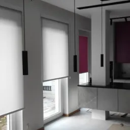 Nowoczesna kuchnia z trzema oknami osłoniętymi jasnymi roletami materiałowymi, uzupełniona minimalistycznymi czarnymi lampami wiszącymi, z widoczną częścią zabudowy kuchennej w odcieniach szarości...