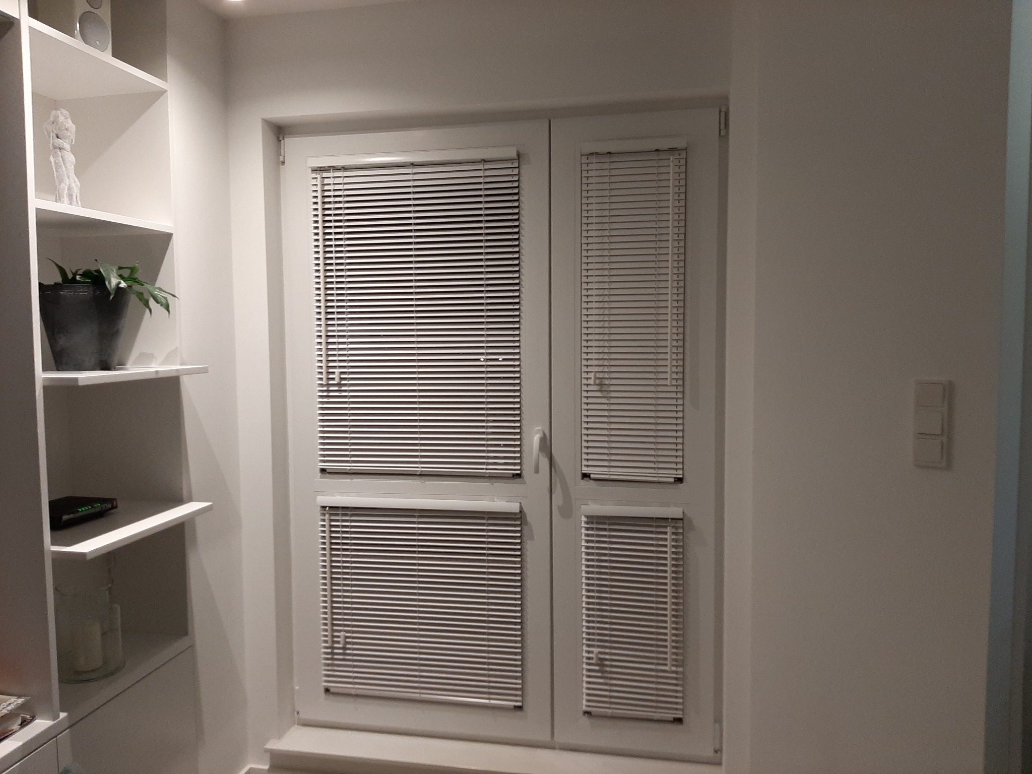 Białe, aluminiowe żaluzje poziome zamontowane na dwuskrzydłowych drzwiach balkonowych, widoczne z boku, w tle białe ściany i regał z dekoracjami.