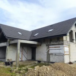 Surowy stan domu z szarą dachówką i oknami dachowymi, widoczne elementy konstrukcyjne, zabezpieczone okna, ziemia przed budynkiem.