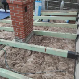 Ocieplenie poddasza wełną mineralną między krokwiami, widoczny komin z czerwonej cegły i fragment dachu z folią paroizolacyjną rozwijaną przez dekarza.