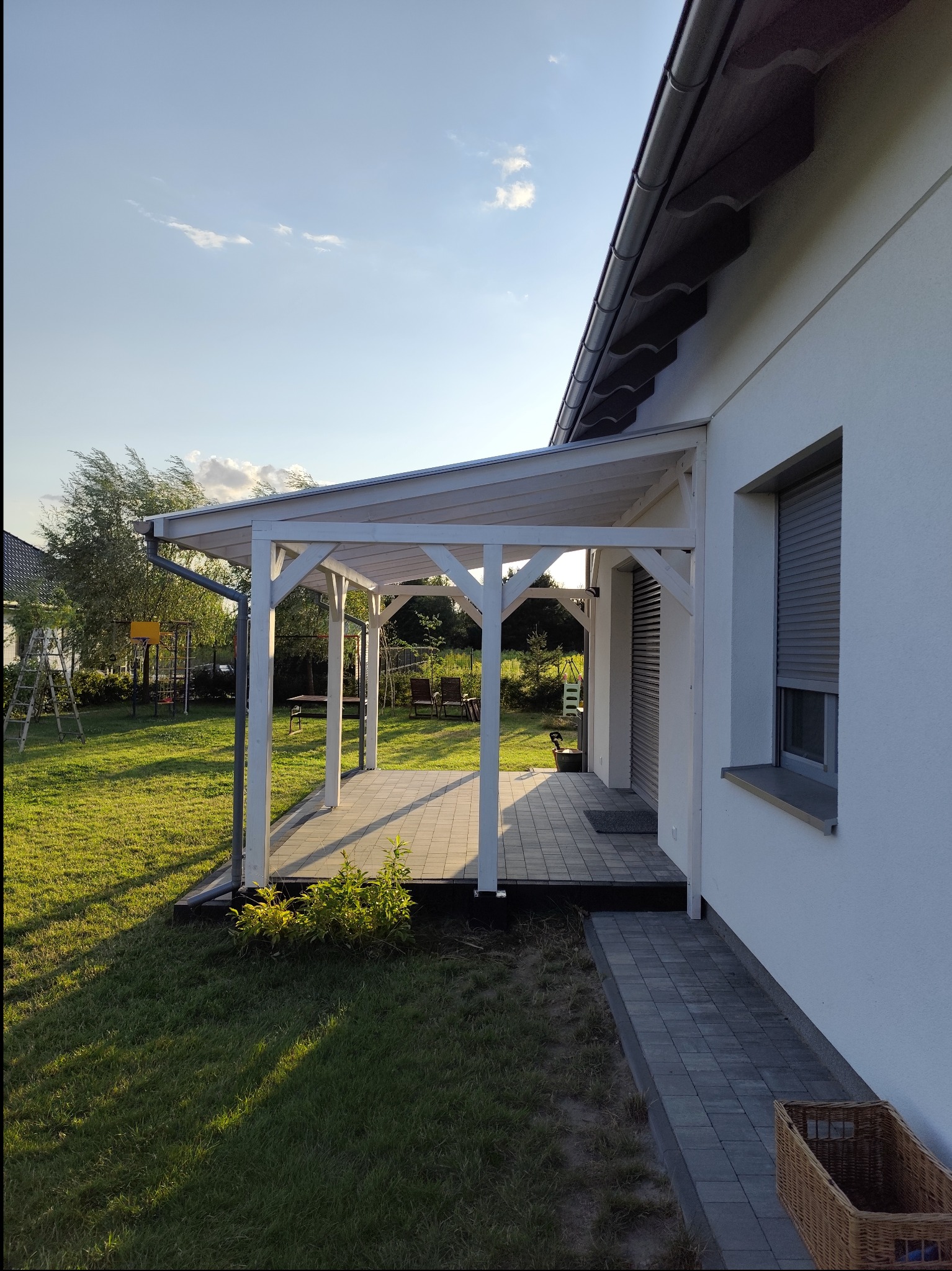 Biała pergola z zadaszeniem przylegająca do ściany domu z roletami, widok na trawnik, plac zabaw i drzewa w tle, kompozycja z perspektywy ziemi.