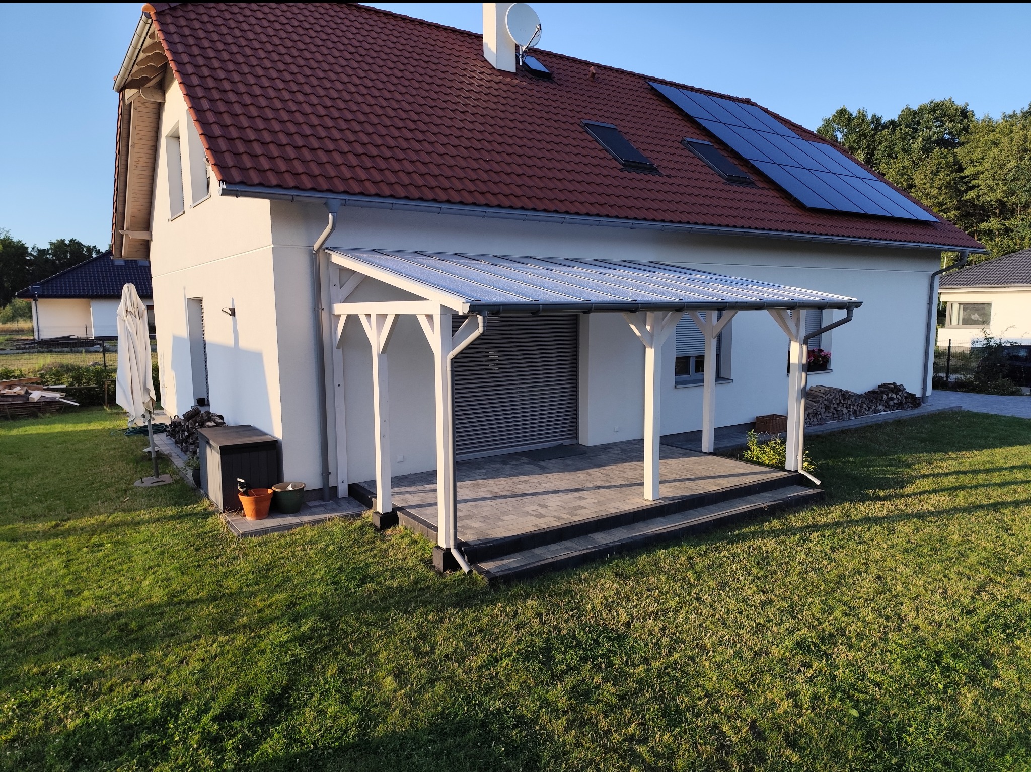 Biała pergola z przezroczystym zadaszeniem przylegająca do jasnego domu z czerwoną dachówką i panelami słonecznymi, widok na zadbany trawnik.