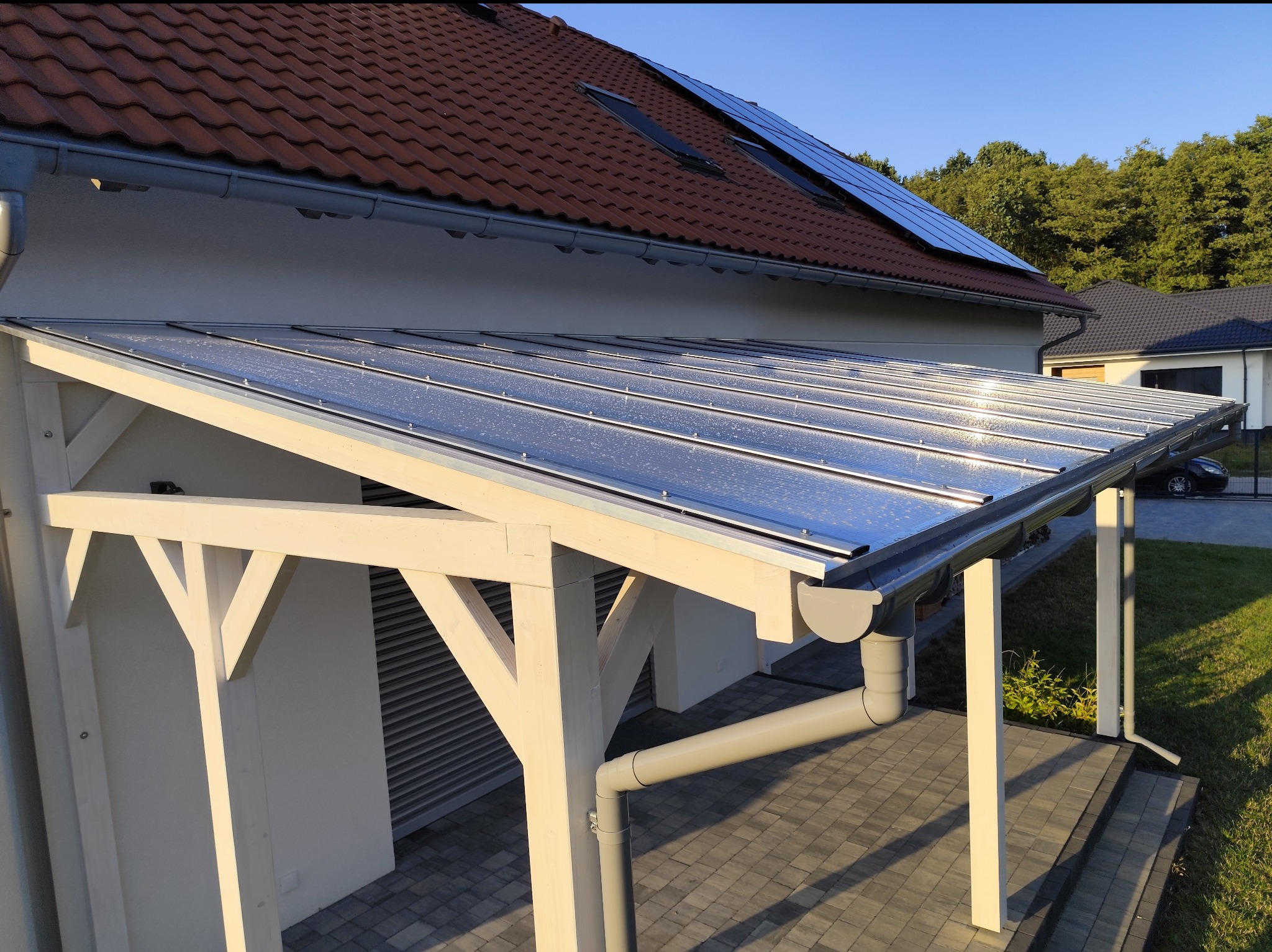 Biała, drewniana konstrukcja z przezroczystym zadaszeniem, przylegająca do domu z brązowym dachem i panelami słonecznymi, widoczna kostka brukowa i trawnik w słoneczny dzień.