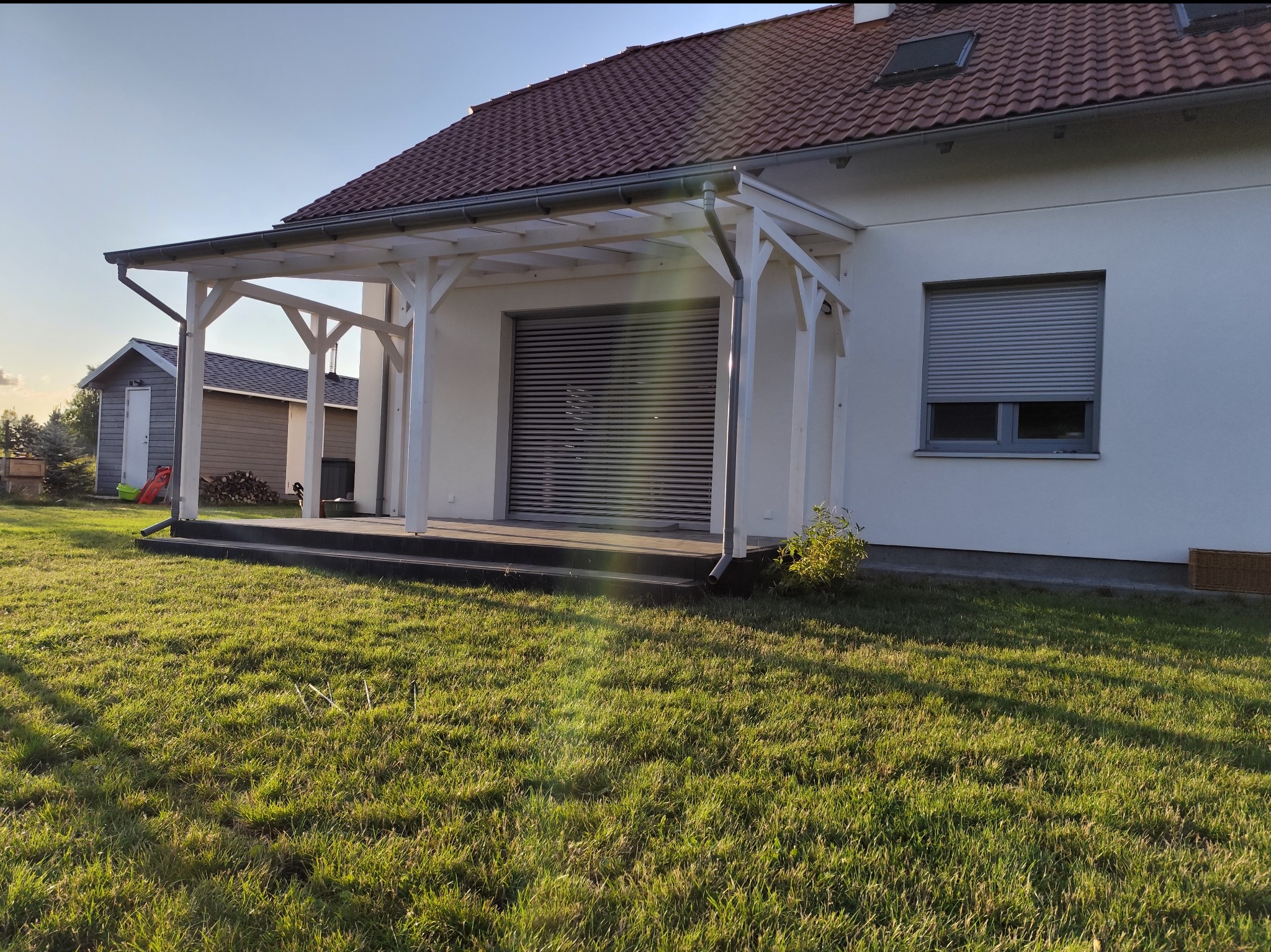Biała konstrukcja zadaszenia przy ścianie domu z roletami, widok na trawnik i budynek gospodarczy w tle.